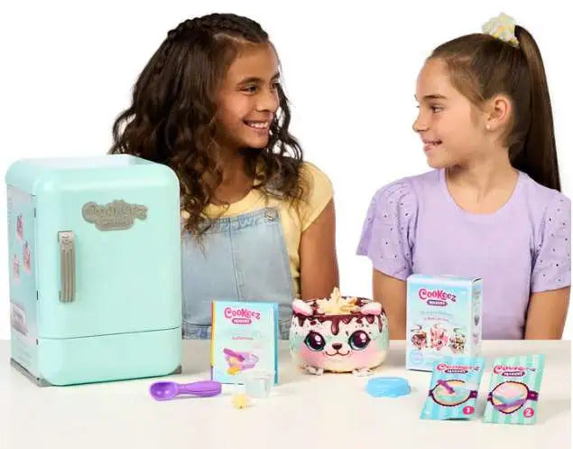 Two children with a toy kitchen set including a mint green refrigerator, cookablez packaging, and a pink toy cake.